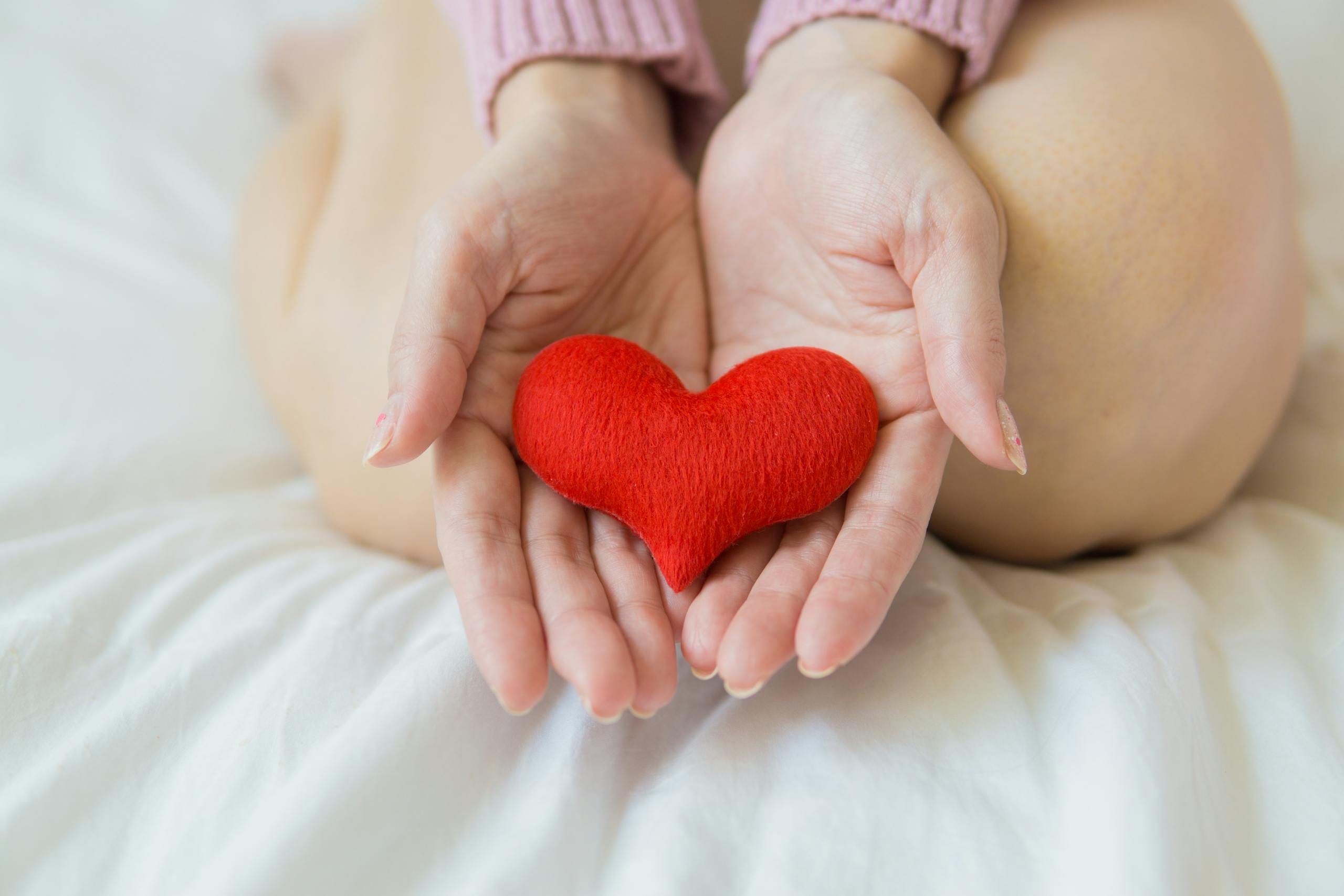 Unrecognizable female sitting with bare legs on white sheet with small red heart in hands in light room in daytime